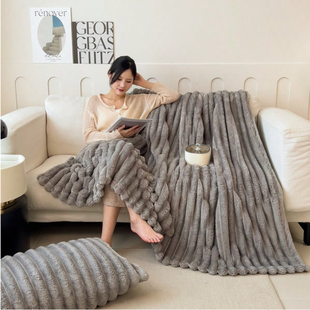 Frau mit Buch unter einer hellgrauen Kuscheldecke aus Kunstfell mit Rippenstruktur auf dem Sofa
